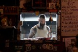 Inside of a dark bar, the only light comes from a small window in the wall leading to the back kitchen. Surrounding the window are stickers and plaques on the wall, layered over time. Inside the window a tall person stands, covered in black and grey tattoos and and looking down over their long beard.