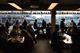 horizontal photo of the inside of a busy bar. outside the clear windows are rows of cars parked as the sun sets