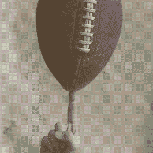 A sepia-toned image of a hand balancing and spinning an American football on one finger against a softly textured, neutral background.