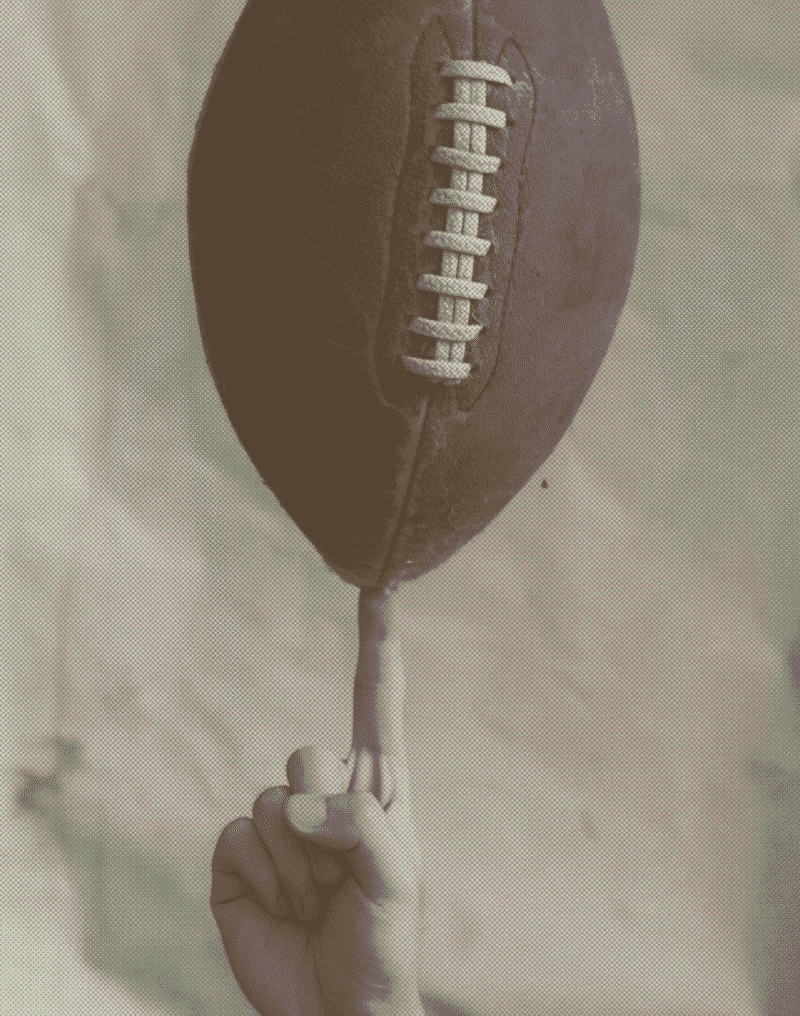 A sepia-toned image of a hand balancing and spinning an American football on one finger against a softly textured, neutral background.