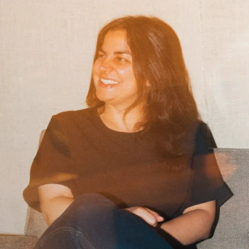 A woman with long dark hair sits on a chair, smiling and looking to the side.
