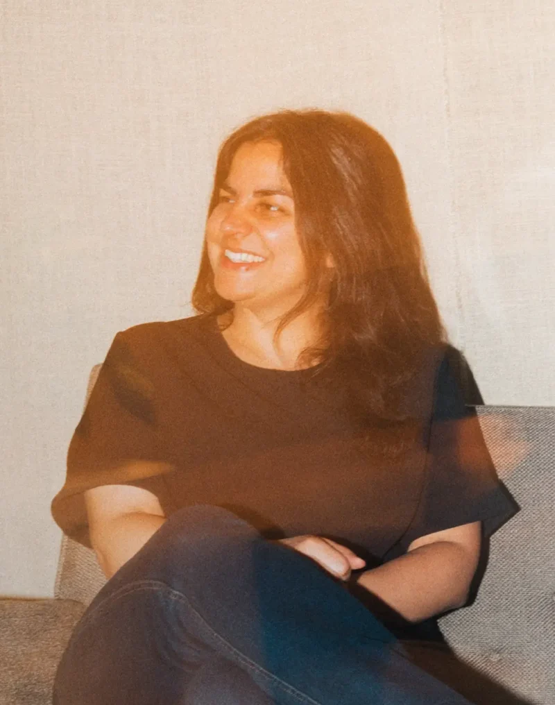 A woman with long dark hair sits on a chair, smiling and looking to the side.