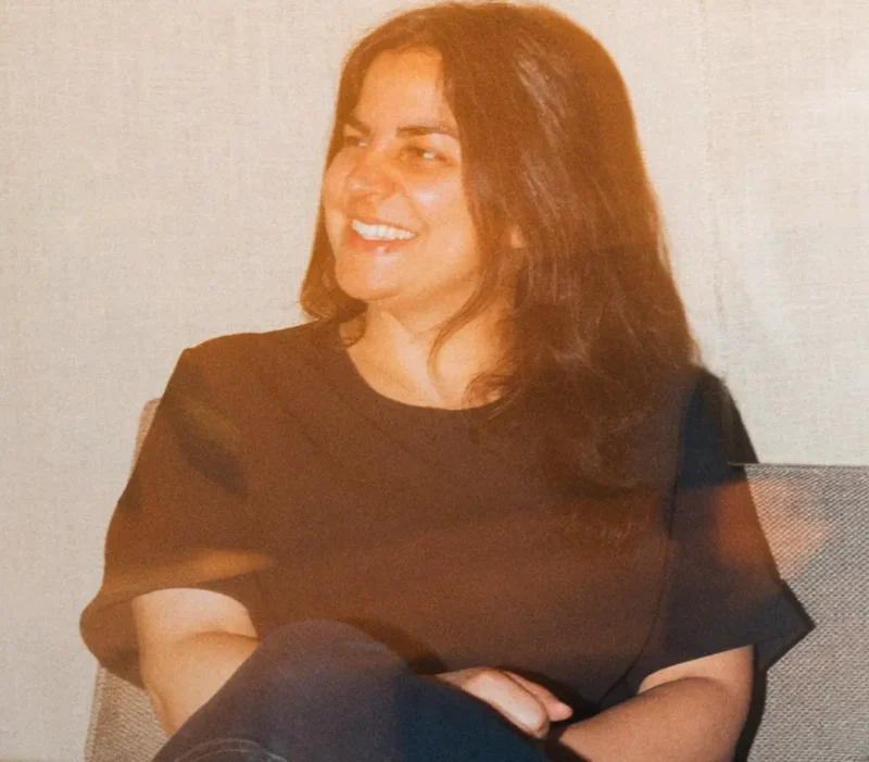 A woman with long dark hair sits on a chair, smiling and looking to the side.